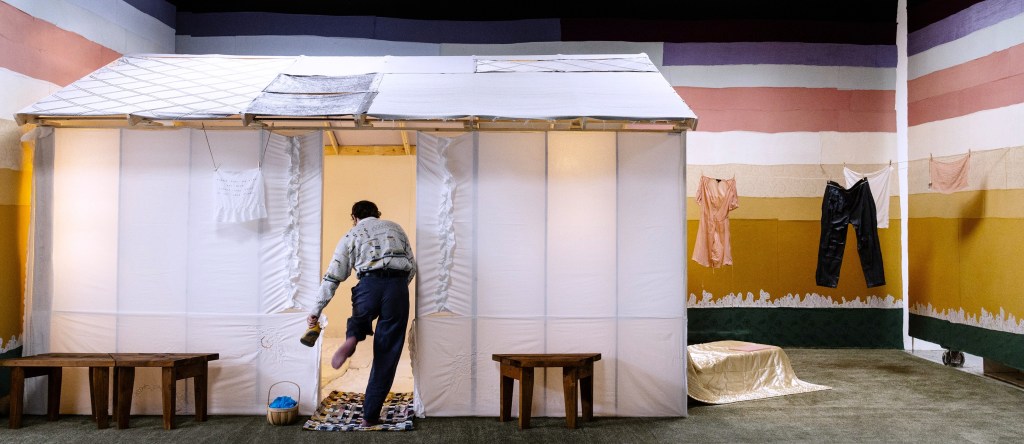 "Copper View," 2025. Installation view of a timber framed cottage with white bed linens for walls and grey linens for roof. Strips of linens cover the walls of the exhibition space mimicking the colors of a sunset. To the right of the cottage is an faux outdoor area with green carpet and a lawn chair covered in a gold quilt. There are clothes hung on a line in the outdoor area. The artist is standing in the doorway of the cottage removing his shoes. 